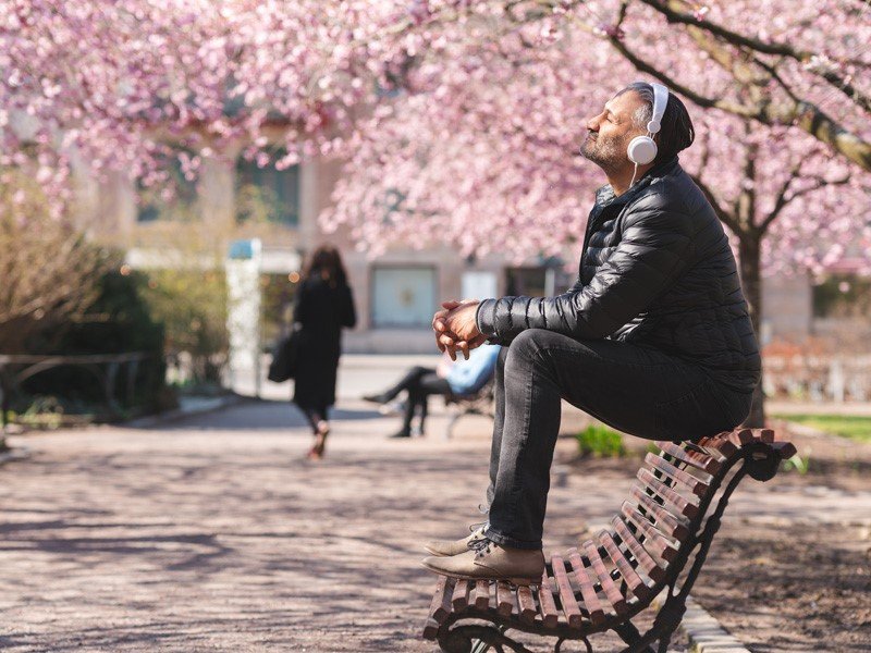 Life Extension, man with headphones sitting on top of a bench in a park druing cherry blossom enjoying the sun